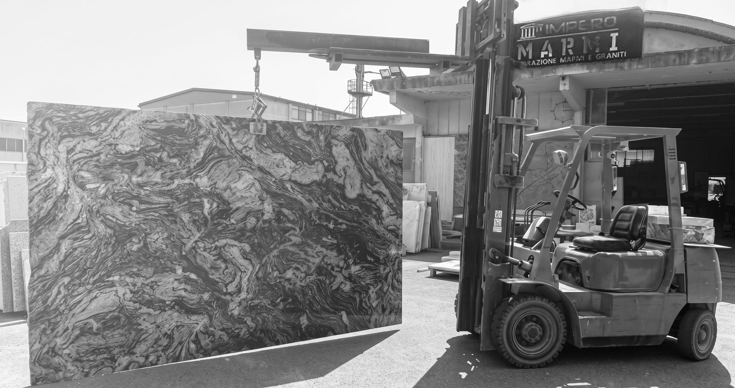 Il piazzale del laboratorio Impero Marmi: un muletto solleva una lastra di granito venato davanti all'insegna, in bianco e nero